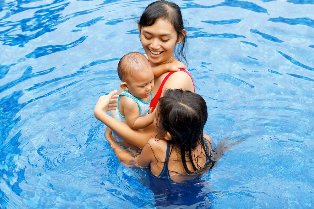 an adult woman, perhaps the mother with a baby and a little girl smiling and standing in the water of a swimming pool