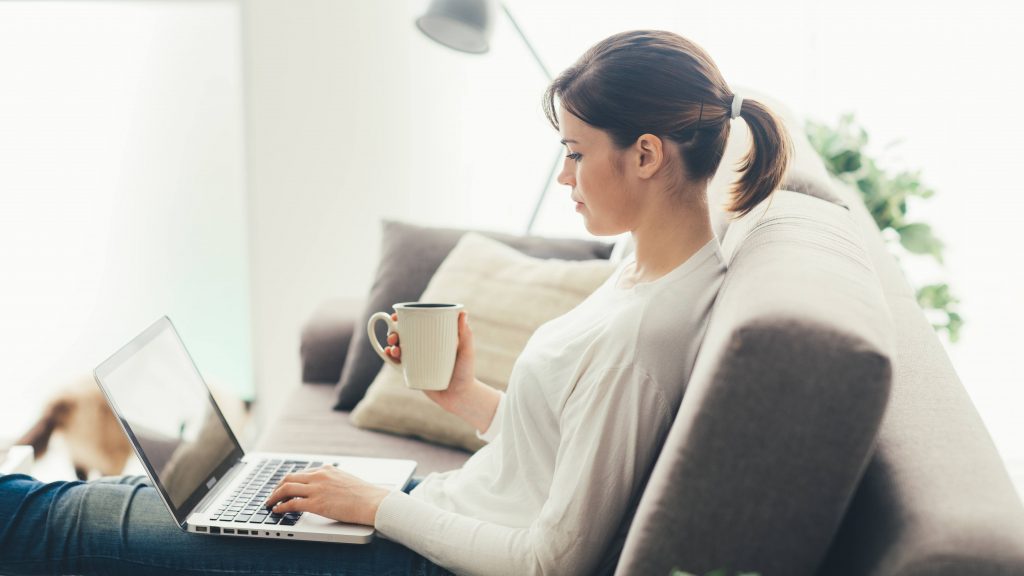 a side view of a young woman sitting on a couch with a cup in her hand, looking at a laptop screen