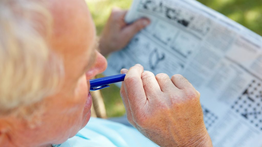 an elderly man, senior citizen sitting outdoors doing the crossword puzzle in the newspaper, perhaps to exercise his brain and his memory, keeping his mind health