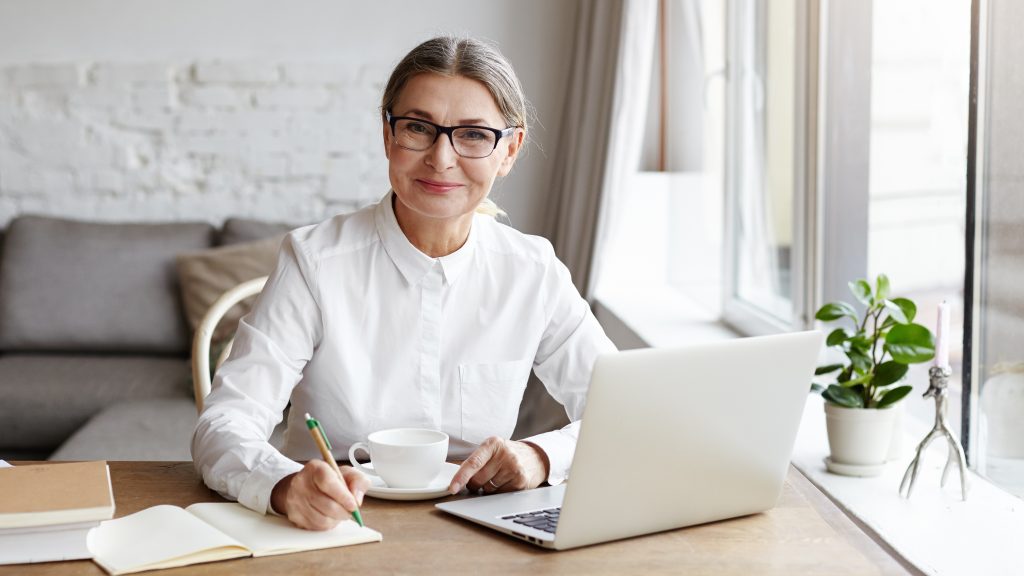 Una mujer mayor sentada al escritorio de una habitación soleada sonríe frente a un computador portátil mientras sostiene una taza y escribe un diario