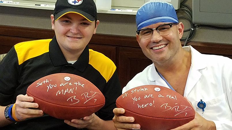 patient Seth Bayles smiling with Dr. John Stulak, each holding autographed footballs from Pittsburgh Steelers quarterback Ben Roethlisberger