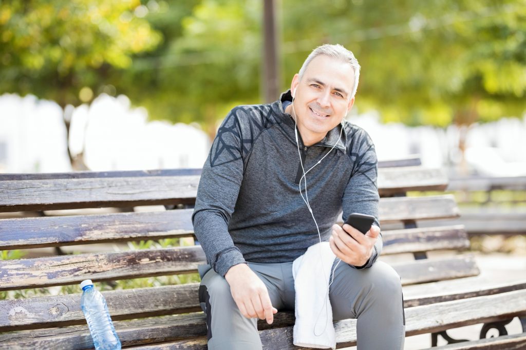 Un hombre maduro y con sonrisa amplia escucha música en el teléfono mientras descansa un poco del ejercicio que está haciendo en el parque