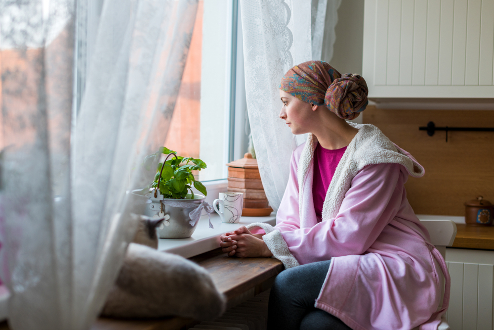 Young adult female cancer patient wearing headscarf and bathrobe, sitting in the kitchen looking out window
