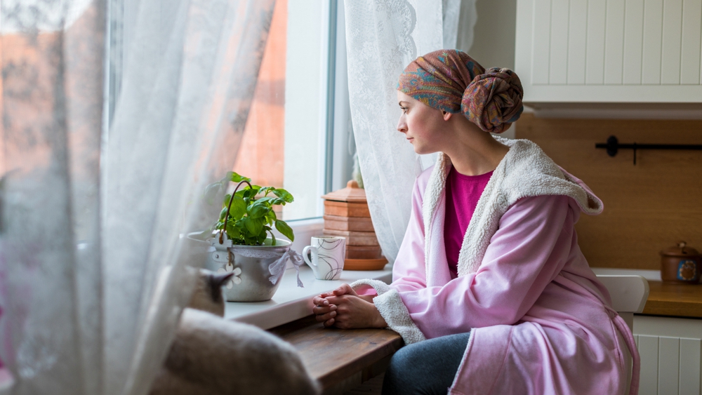 Una mujer joven con cáncer lleva un pañuelo en la cabeza y una bata en el cuerpo mientras está sentada en la cocina y mira por la ventana