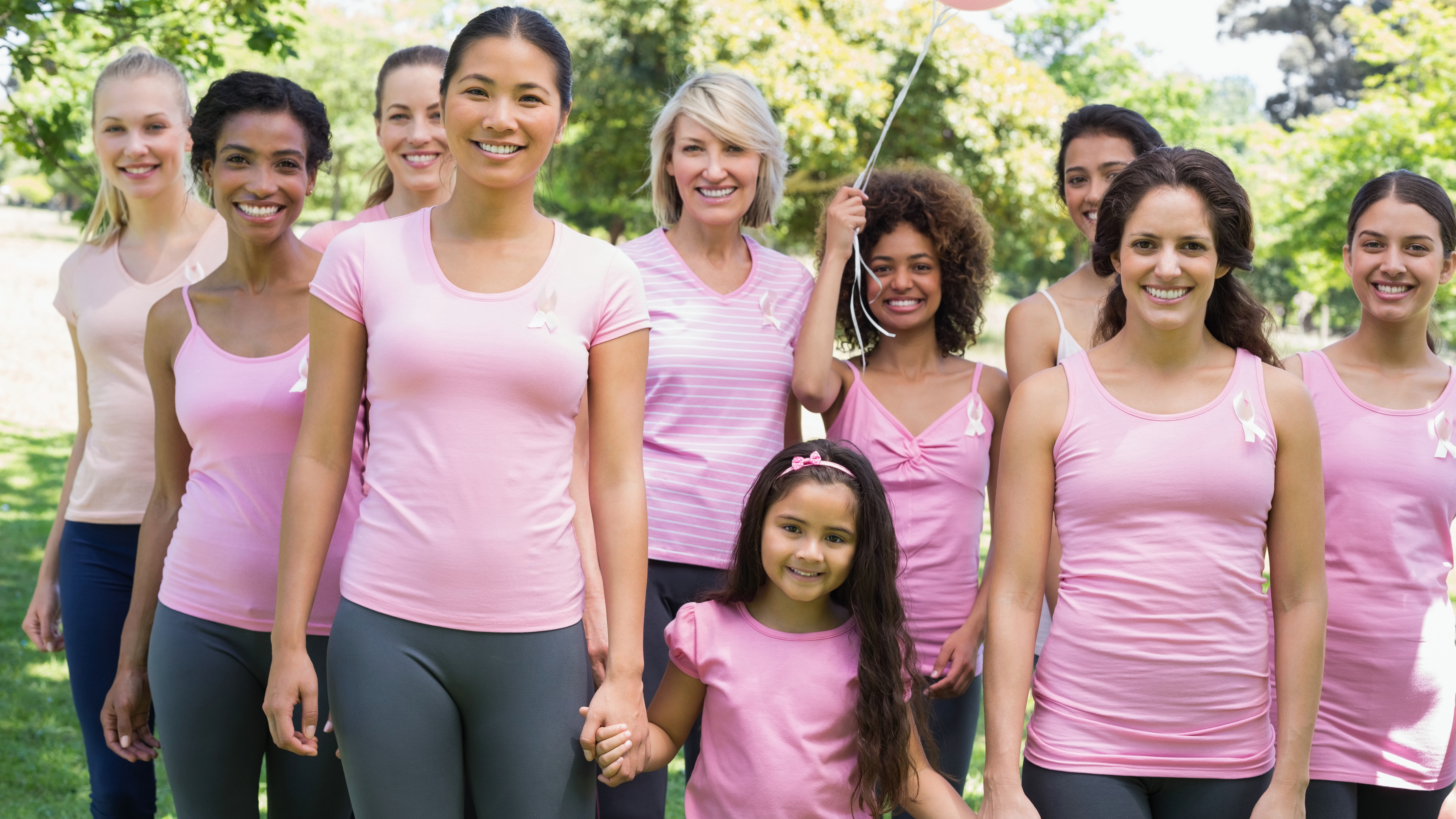 a group of multiethnic women and a young girl standing outside in a park, all wearing pink and supporting breast cancer awareness