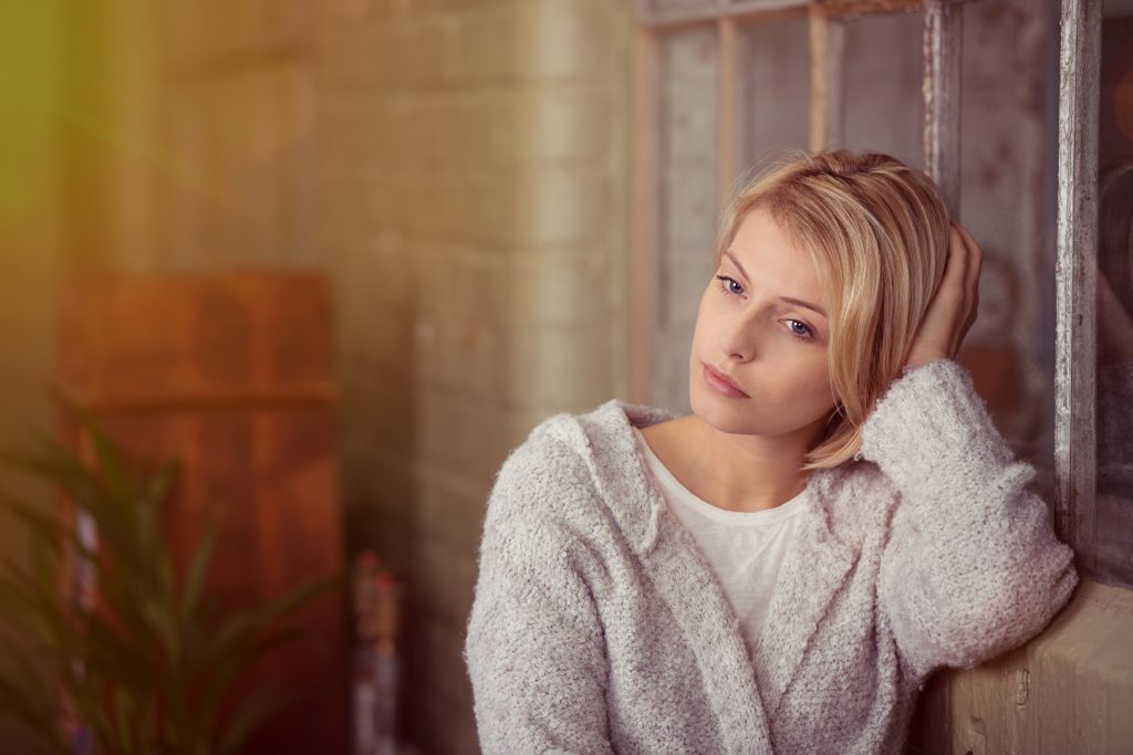 a young woman leaning against a brick wall and window sill, looking serious or sad