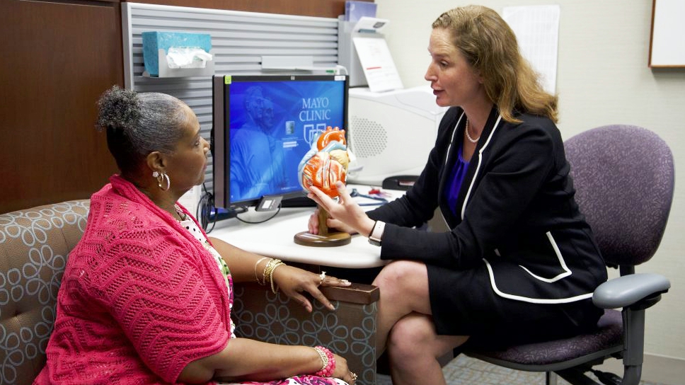 a female cardiologist talking with a woman patient in an exam room about heart disease using a model of a heart