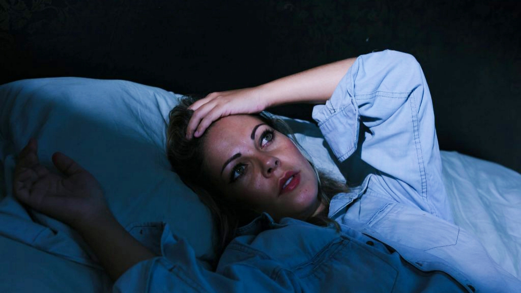 a young woman in bed in a dark room, lying awake with her eyes open and her hand on her forehead, not sleeping and suffering from insomnia or stress