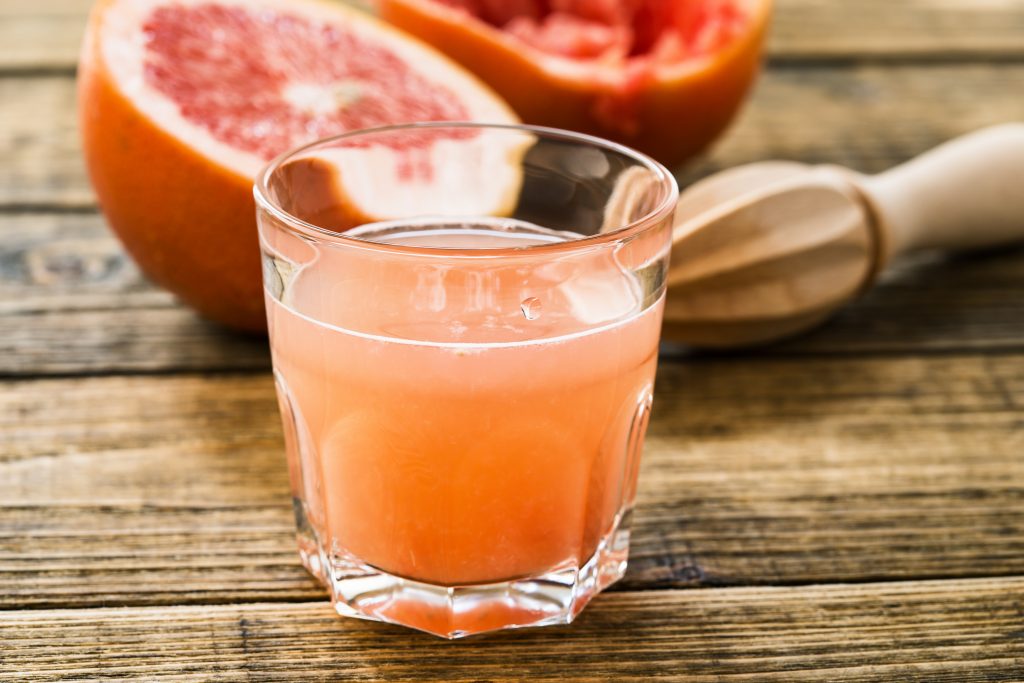 a glass of fresh pink grapefruit juice on a rough wooden surface, with two grapefruit halves in the background