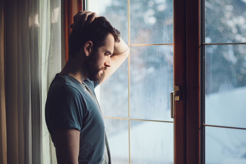 a young man standing by a rain-streaked window, looking sad, worried, depressed