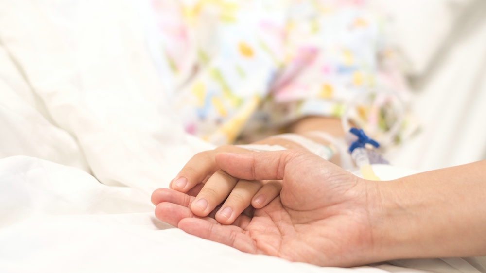 a child patient with IV line in hand sleep on hospital bed with an adult holding her hand for comfort