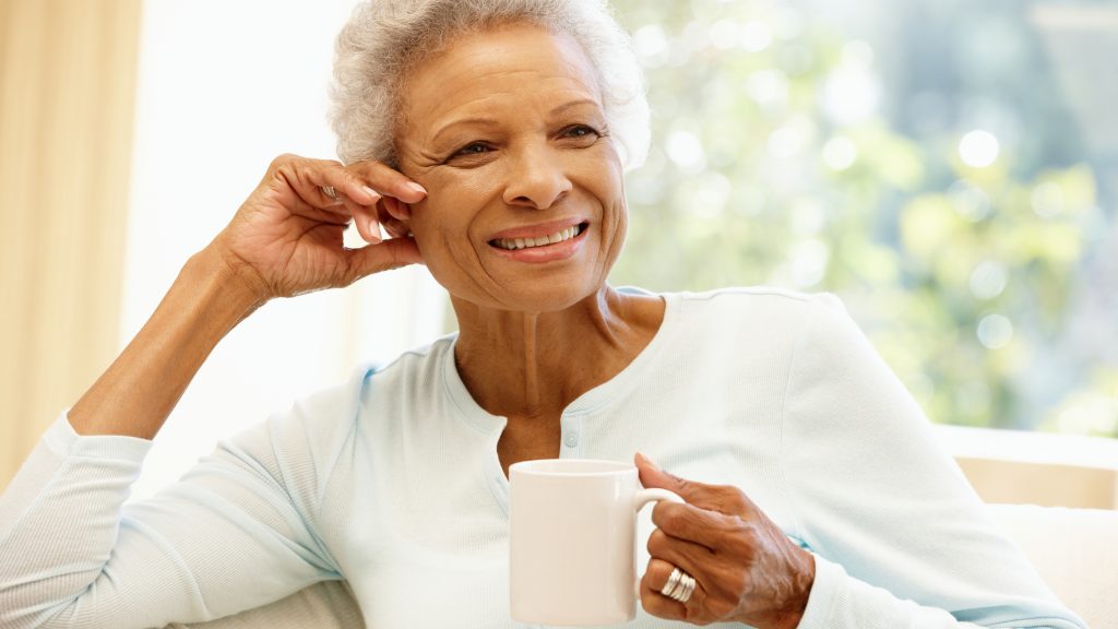 a mature, smiling, confident African American woman smiling and holding a cup of coffee while sitting on a couch near a window