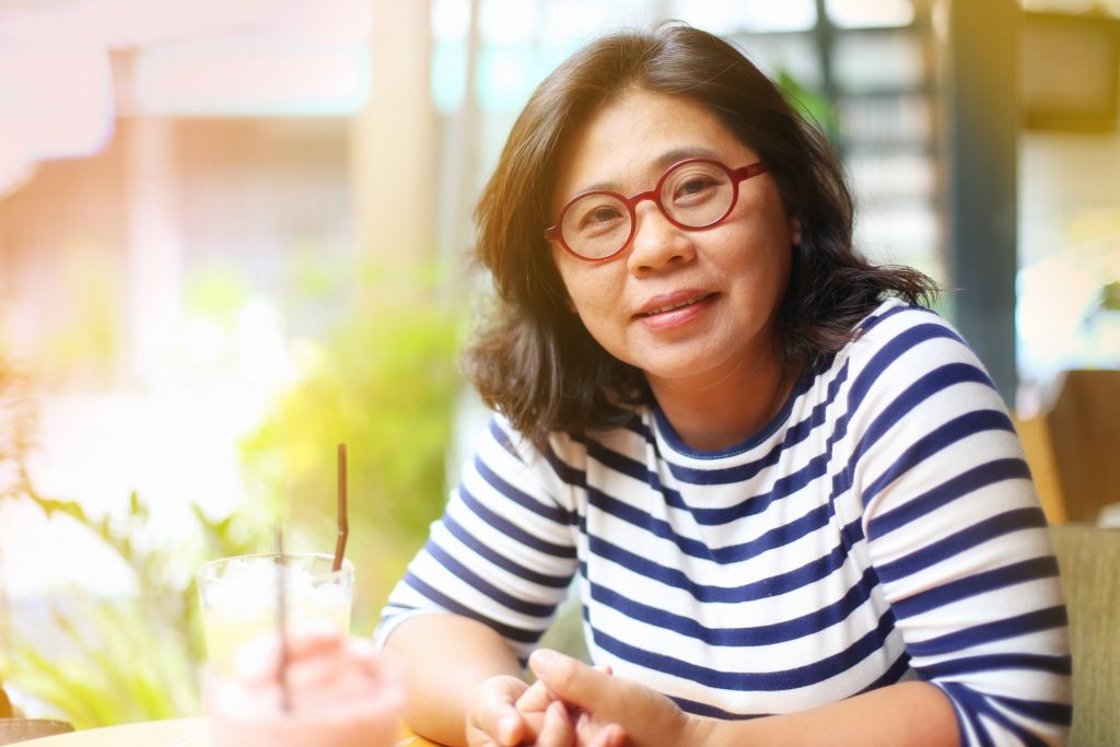 a mature or middle-aged Asian woman wearing glassed and sitting outdoors smiling, looking content and happy
