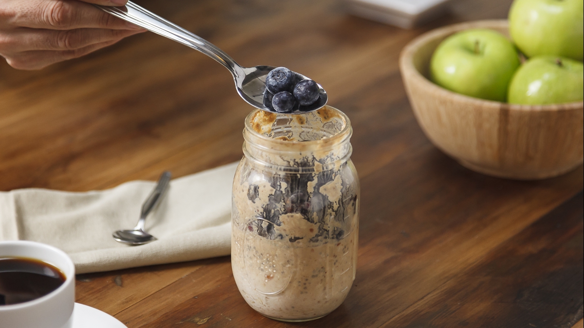 a person putting a spoonful of blueberries into a clear mason jar with refrigerator oatmeal
