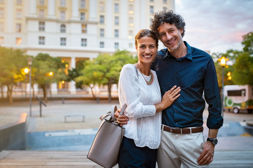 a smiling middle-aged couple, standing together in an outdoor city plaza