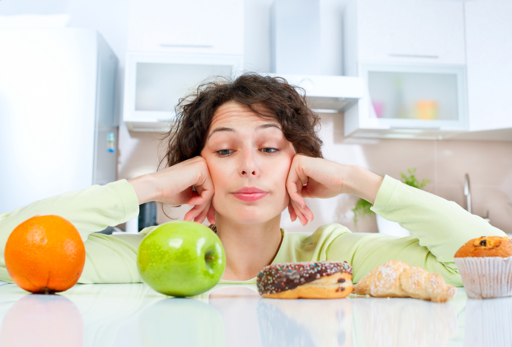 a woman in a kitchen looking at healthy fruit and sugary donuts and pastries, trying to decide which to eat