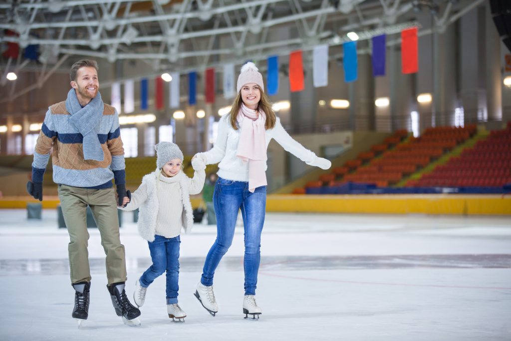 a smiling young family holding hands and ice skating at an indoor rink