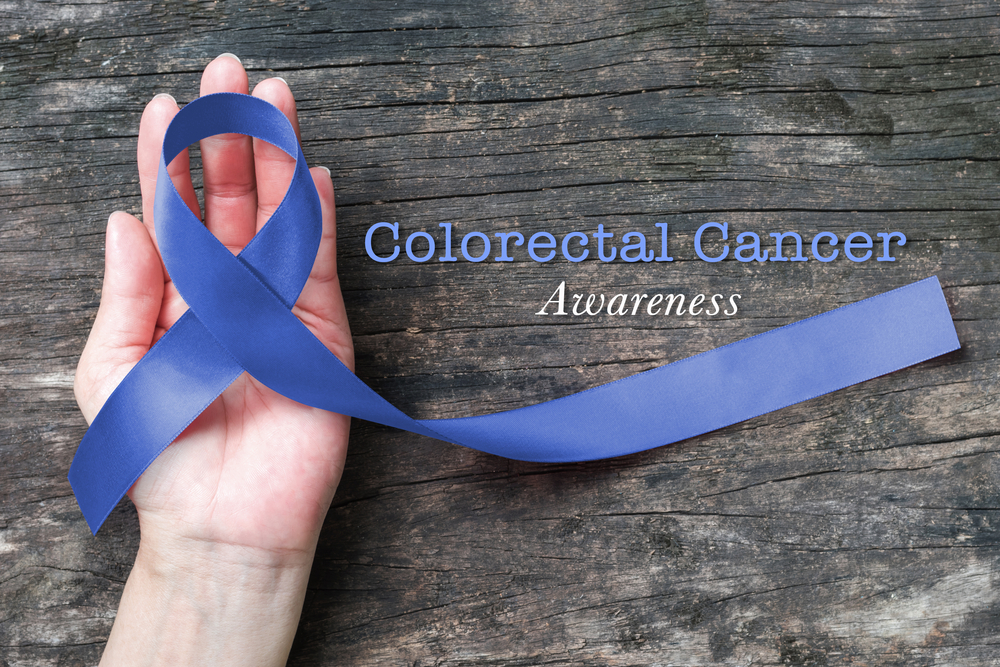 a Caucasion hand palm up on a wooden surface with a dark blue ribbon looped over it and the words 'Colorectal Cancer Awareness' to the side