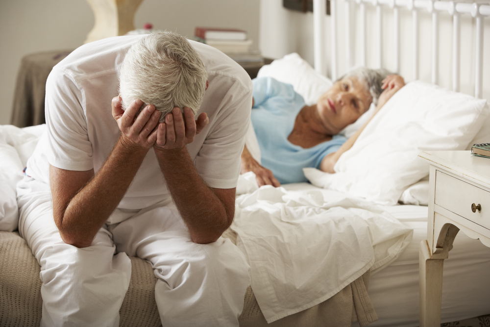an older man sitting on the side of a bed with his head in his hands, with an older woman awake in bed beside him
