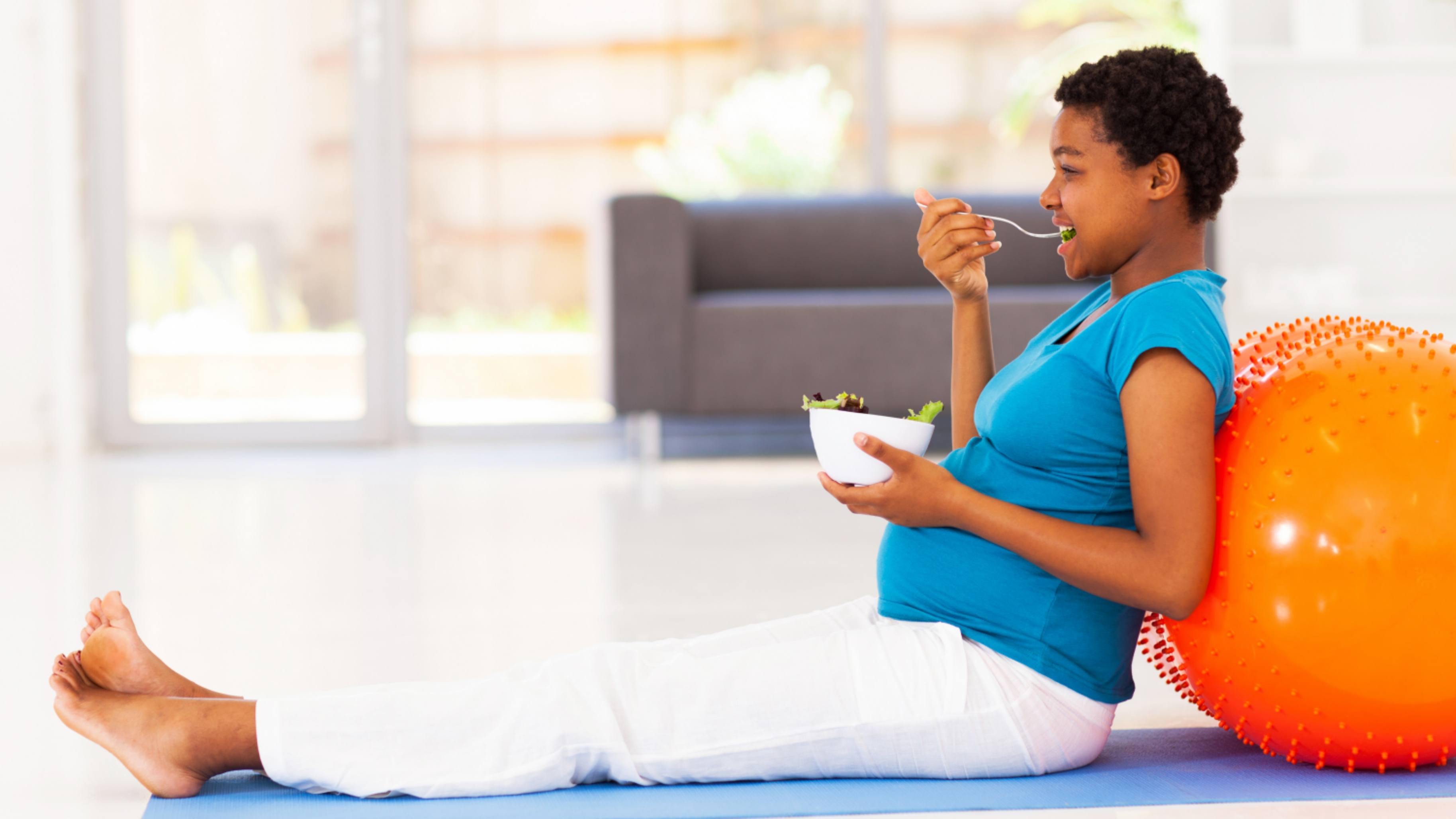 a young African American pregnant woman sitting on a floor with an exercise ball and eating a healthy salad