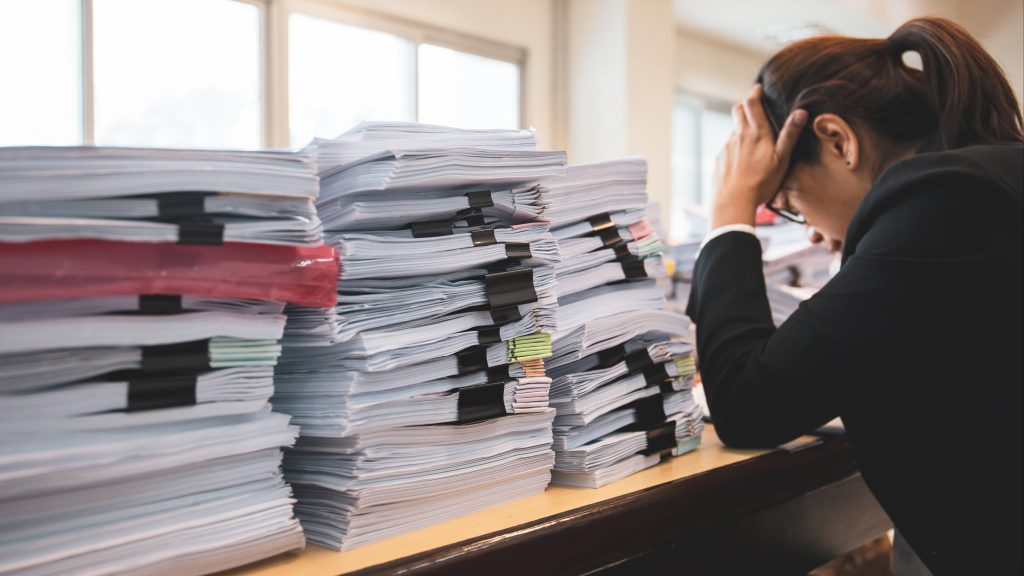 a young business woman looking stressed, holding her head in her hands at a work desk piled high with paperwork