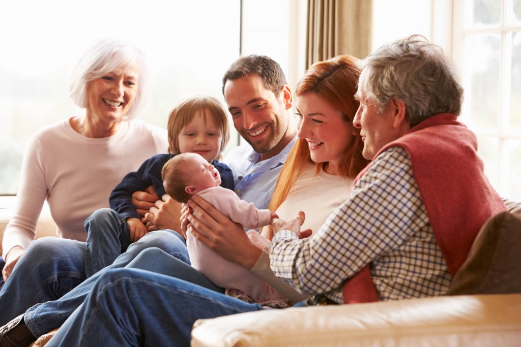 a happy, smiling multigenerational family sitting on a sofa, including grandparents, parents, young child and baby