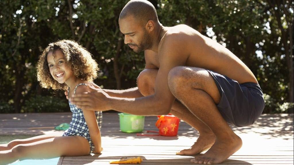 a young girl sitting on the side of a swimming pool with an adult man, perhaps her father, putting sunscreen on her
