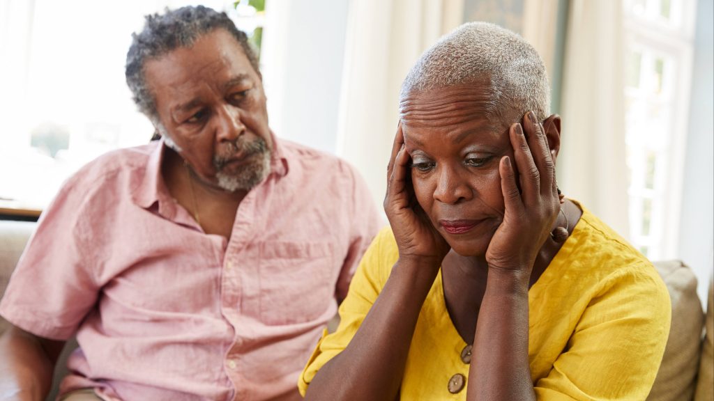 a sad and worried looking African Americanmiddle-aged couple sitting on a couch, with the woman holding her head