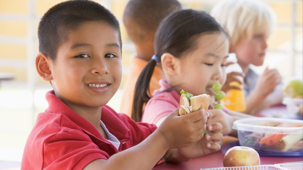 children eating at a school lunch table, a little boy holding a sandwich and a little girl eating grapes