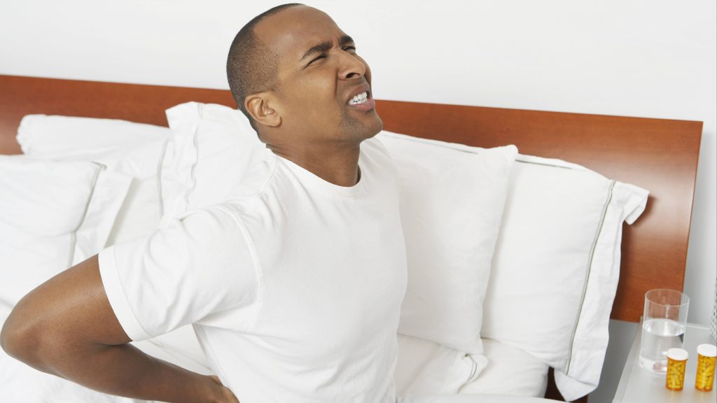 a young African American man sitting on the edge of a bed suffering with back pain