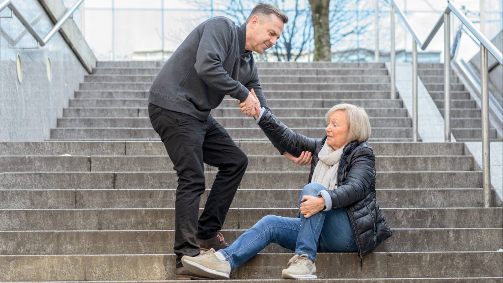 a senior or older woman having fallen down some stairs, holding her knee and possibly injured, and being helped up by a man, a Good Samaritan