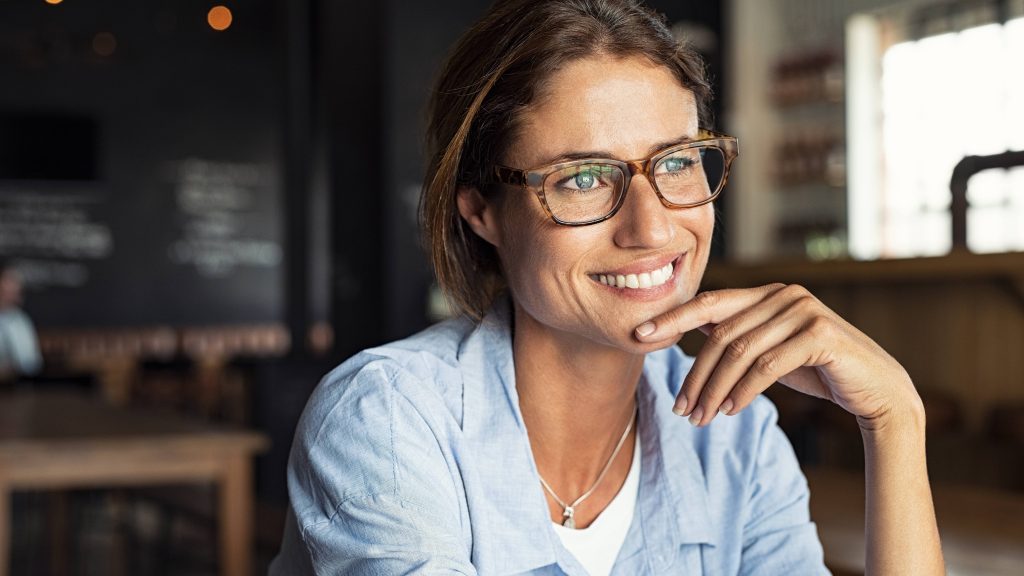 a close-up of a smiling middle-aged woman in glasses, sitting in a coffee shop