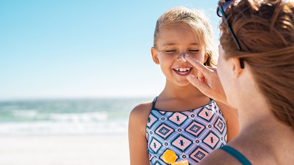 a woman applying sunscreen to the nose of a laughing young girl at the beach