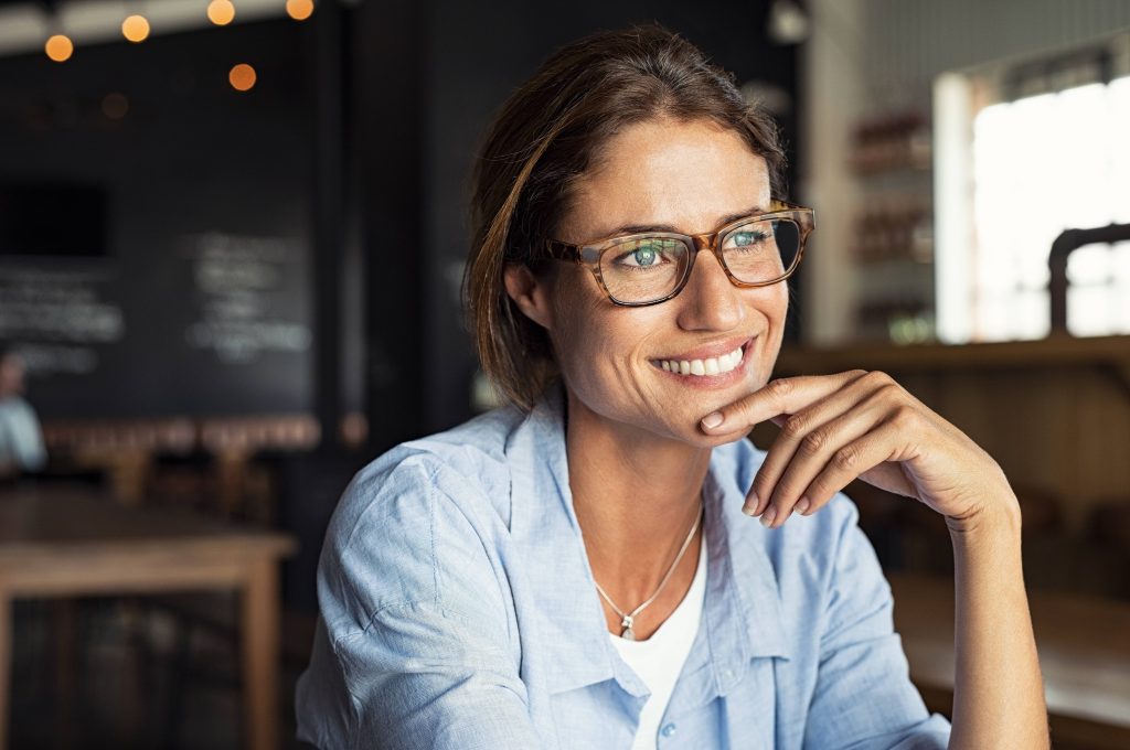 a close-up of a smiling middle-aged woman in glasses, sitting in a coffee shop