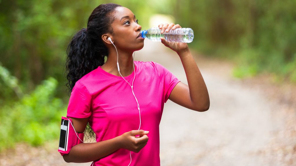 a young African American woman jogging, running, exercising and drinking from a water bottle to stay hydrated