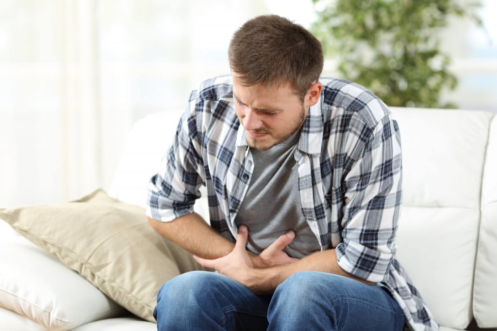a young man sitting on a couch in agony and pain, holding is stomach because of digestive problems or stomach ache