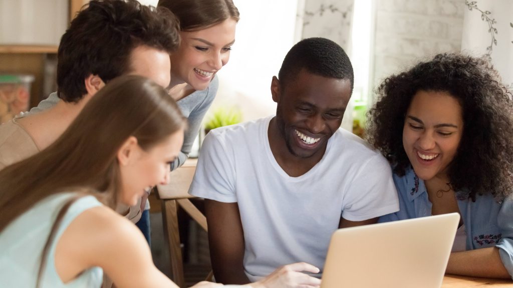 a group of young adult, perhaps university or college students gathered around a laptop computer, smiling, laughing and having fun together