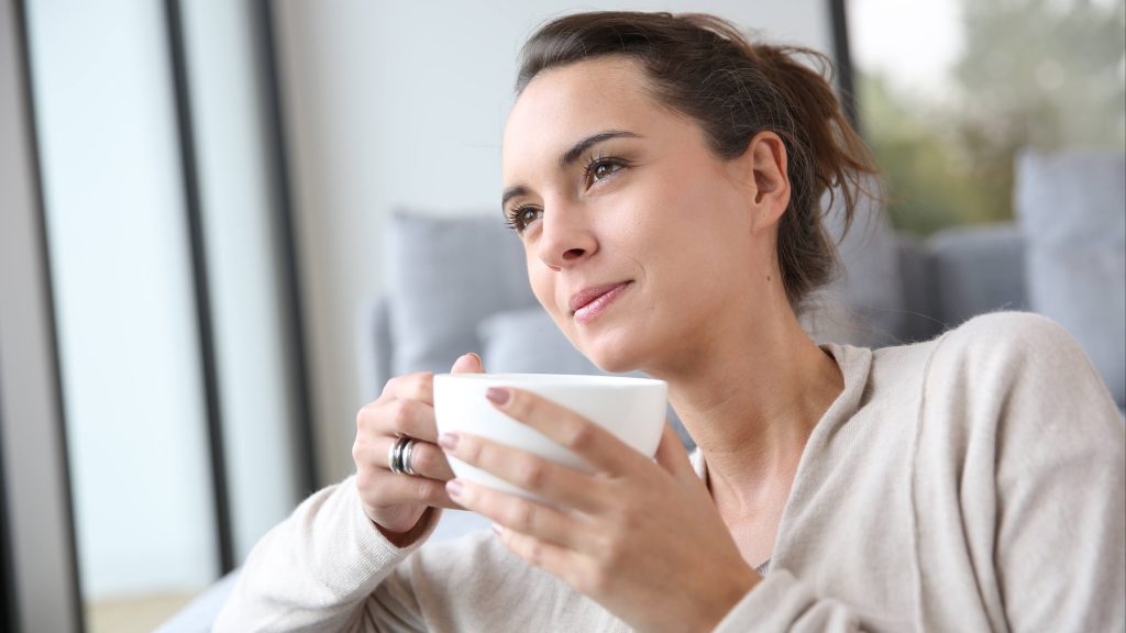 a young woman smiling and looking relaxed, holding a warm cup of tea or coffee, sitting on a couch and looking out a window, caffeine