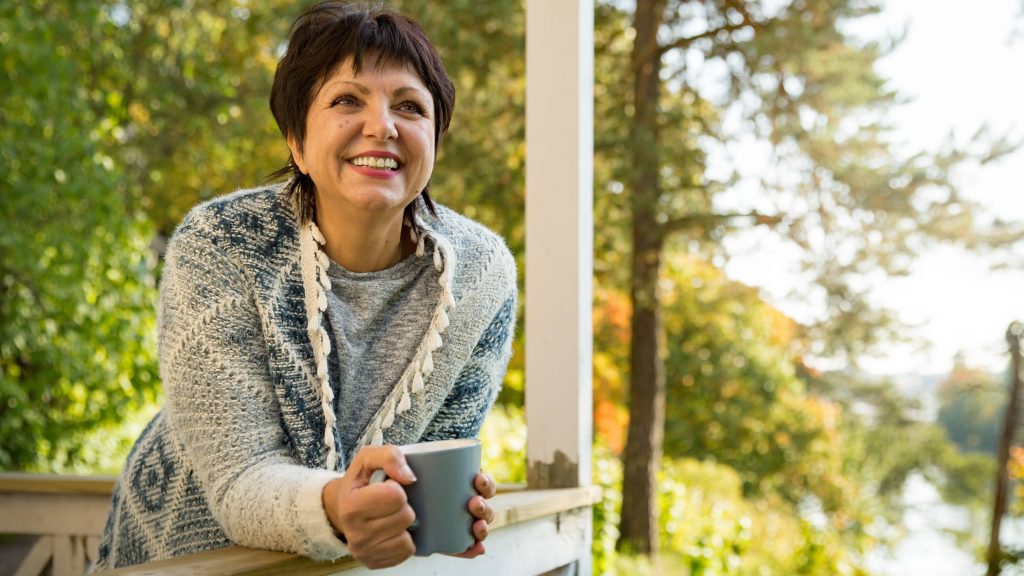 a middle-aged woman in a sweater, smiling and leaning over a porch or balcony railing, holding a cup of coffee