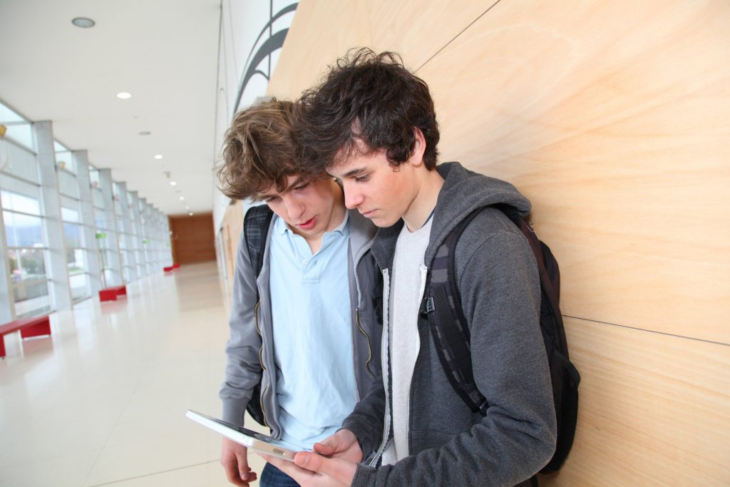 teenage boys looking at a hand-held mobile device, an electronic tablet while standing in a school hallway