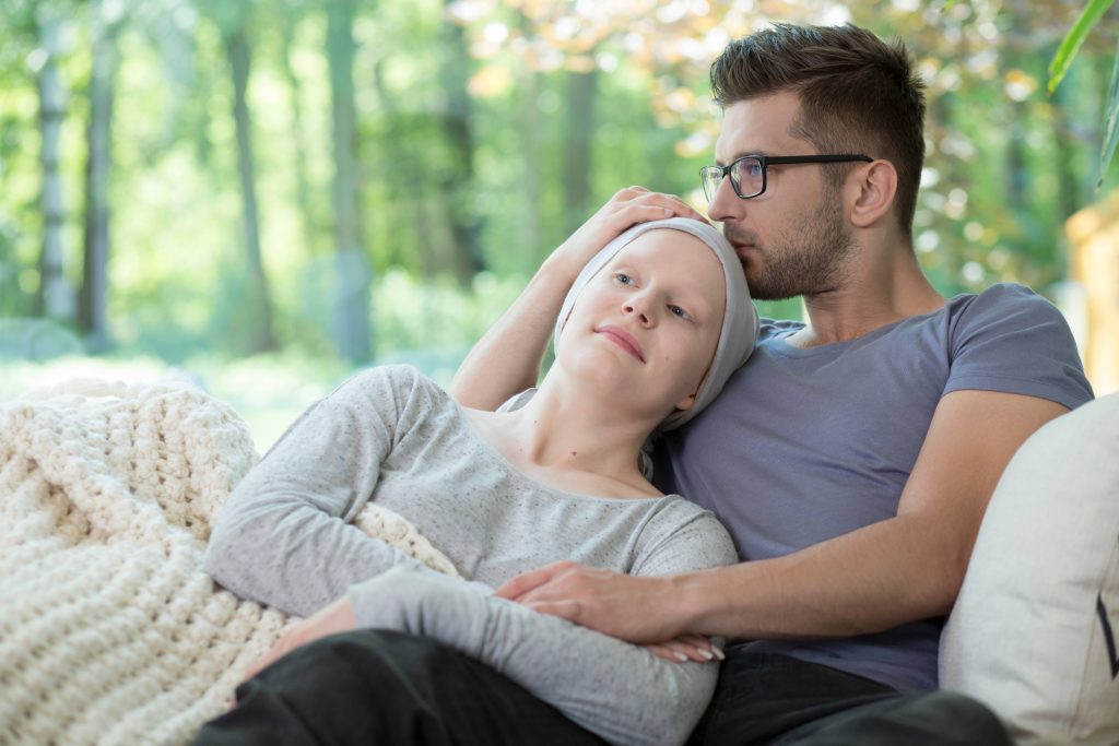 a young couple sitting on a couch or bed with a blanket, comforting each other, perhaps after the woman has had chemotherapy treatment for cancer.