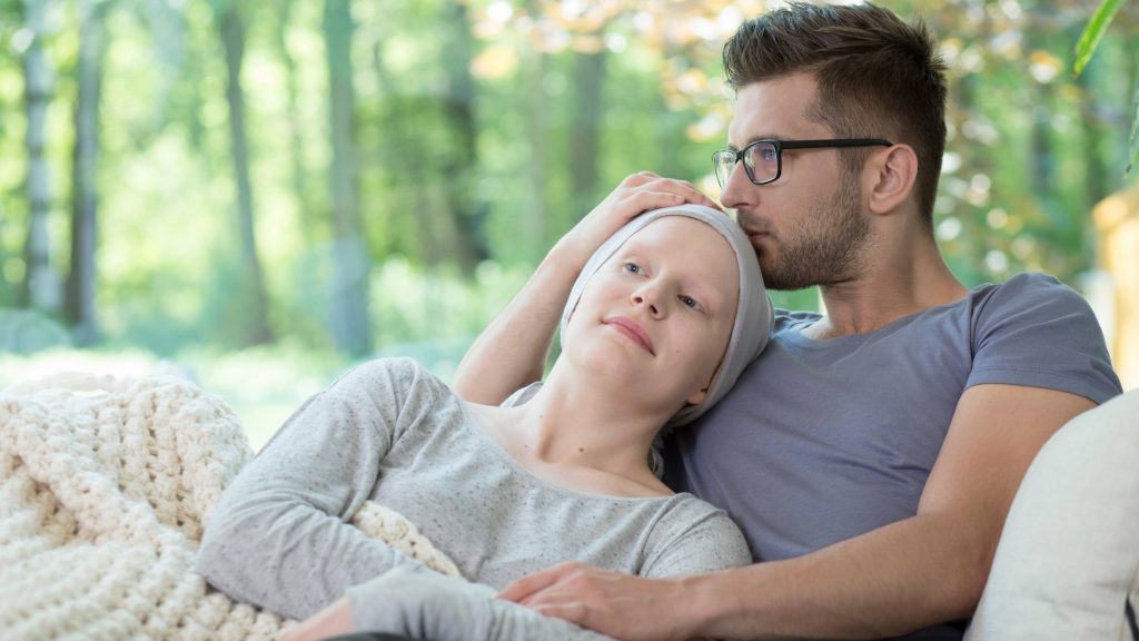 una pareja joven sentada en un sofá o cama con una manta, consolándose mutuamente, quizás después de que la mujer haya recibido un tratamiento de quimioterapia para el cáncer.
