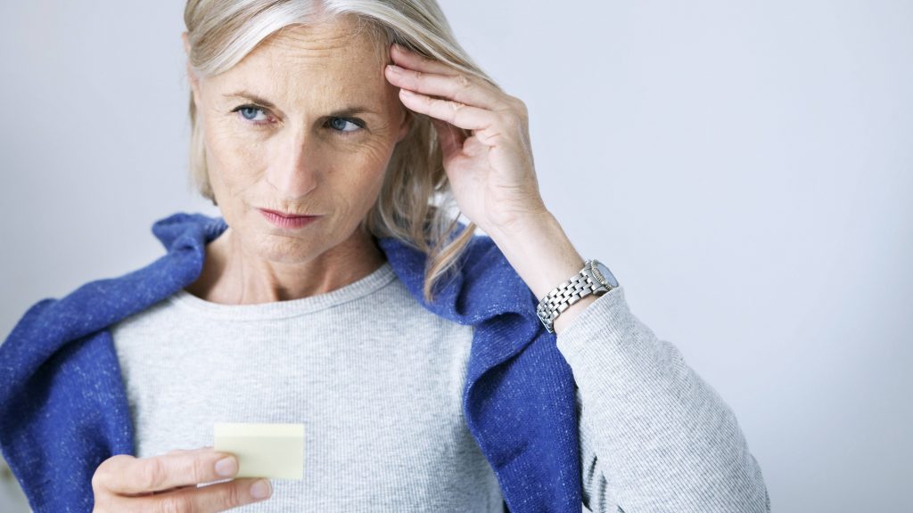 a middle-aged woman looking concerned and holding a small piece of paper, perhaps a list or reminder note, with her hand to her head trying to remember something