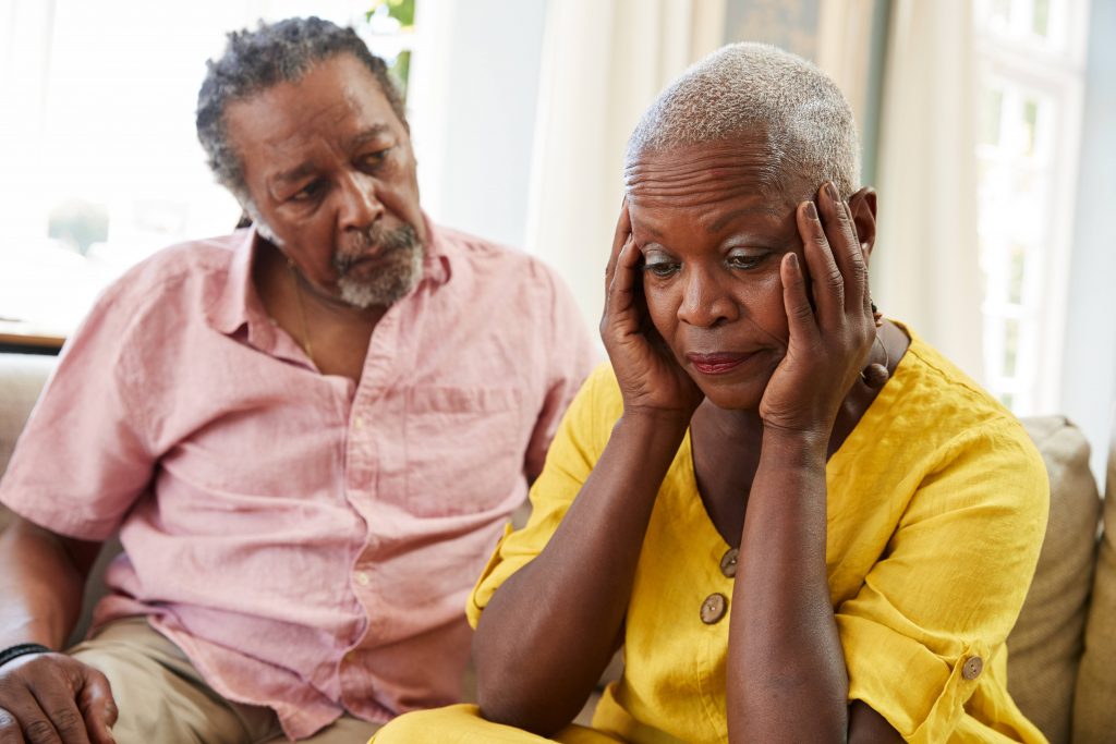 an older couple, a man and a woman, sitting on a couch together looking worried, concerned, sad as the woman holds her head in her hands perhaps having some depression or memory and cognitive function problems
