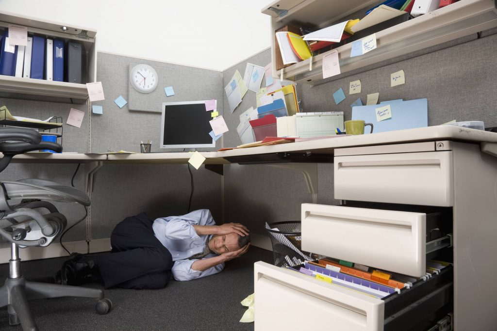 a man grimacing and curled up on the floor under a messy, cluttered desk