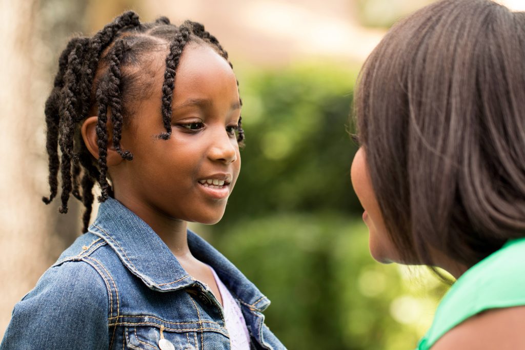 a young African American girl in a denim jacket, smiling and listening to an African American woman, perhaps her mother