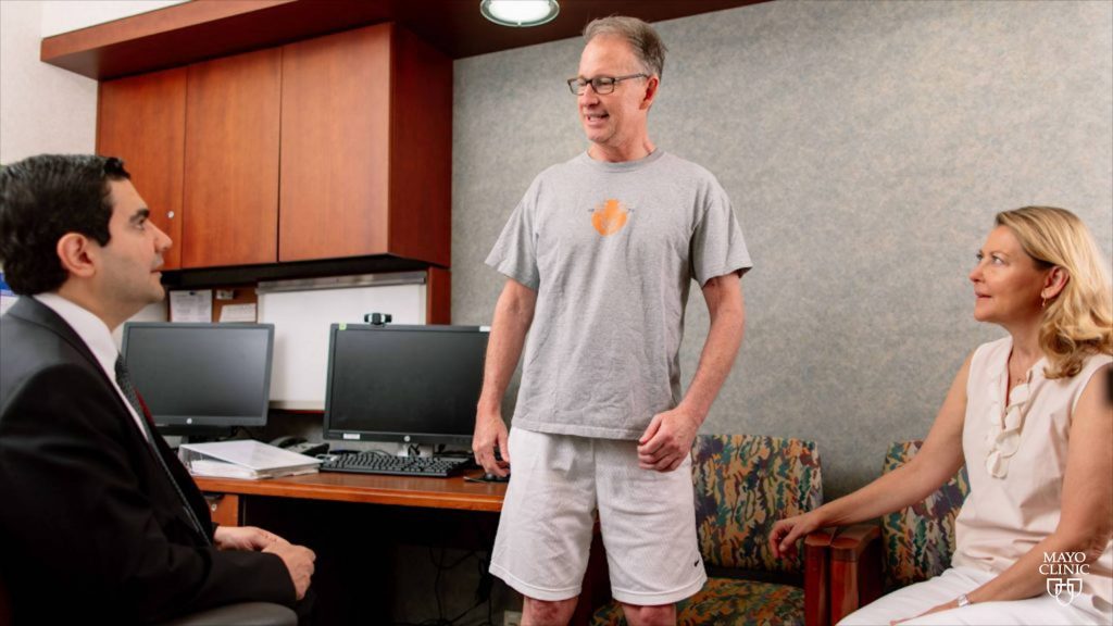 Chris Barr, paciente del ensayo sobre células madre, vestido con pantalones cortos y camiseta aparece sonriente y de pie en la sala de exámenes del Dr. Mohamad Bydon, ante la mirada atenta de su esposa Debbie Barr.