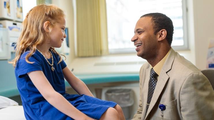 Waleed Gibreel, M.B.B.S., in a business suit smiling in exam room with a little Caucasian girl, pediatric patient, in a blue dress