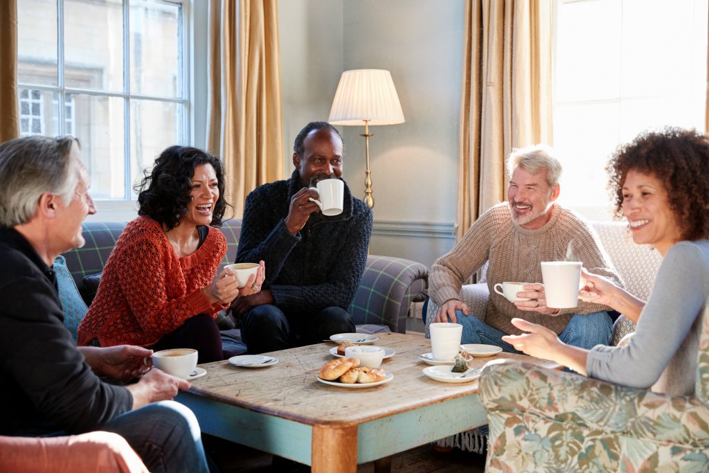 a group of smiling middle-aged men and women sitting around a low table in a living room, enjoying coffee, tea and pastries
