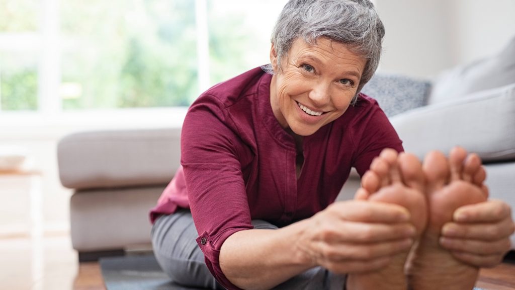 a smiling older woman seated on a yoga mat, doing stretches